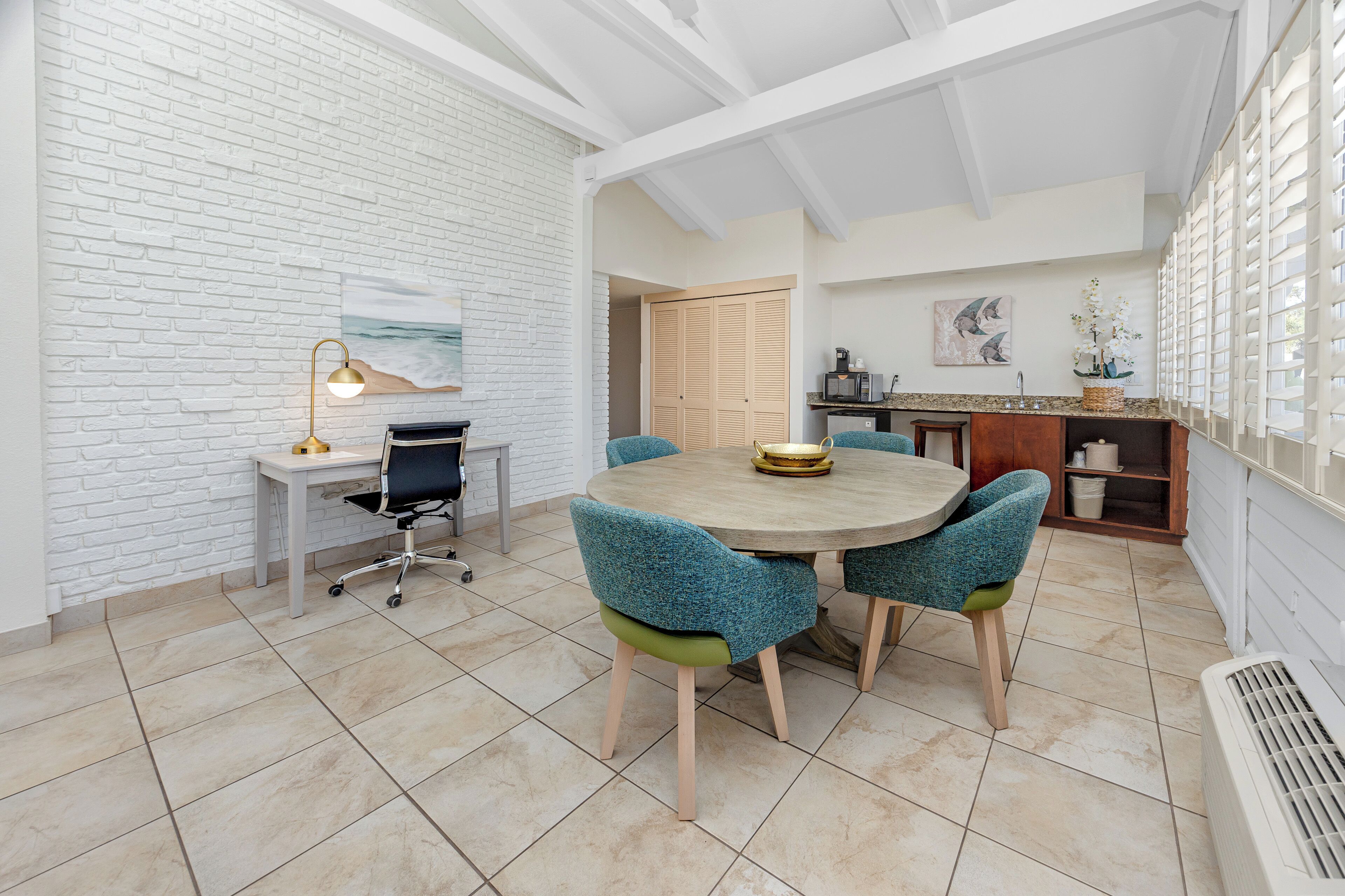 A dining area with a round table and chairs, a white brick wall, and a kitchen area with a microwave and sink.