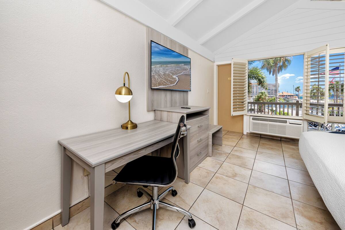 A hotel room with a desk, chair, and a large window with a view of the beach and palm trees.