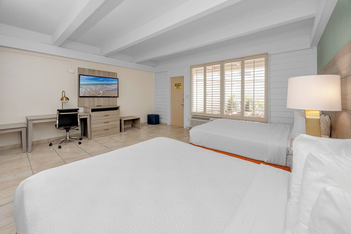 A hotel room with two beds, a desk, a chair, a TV, and a window with blinds.
