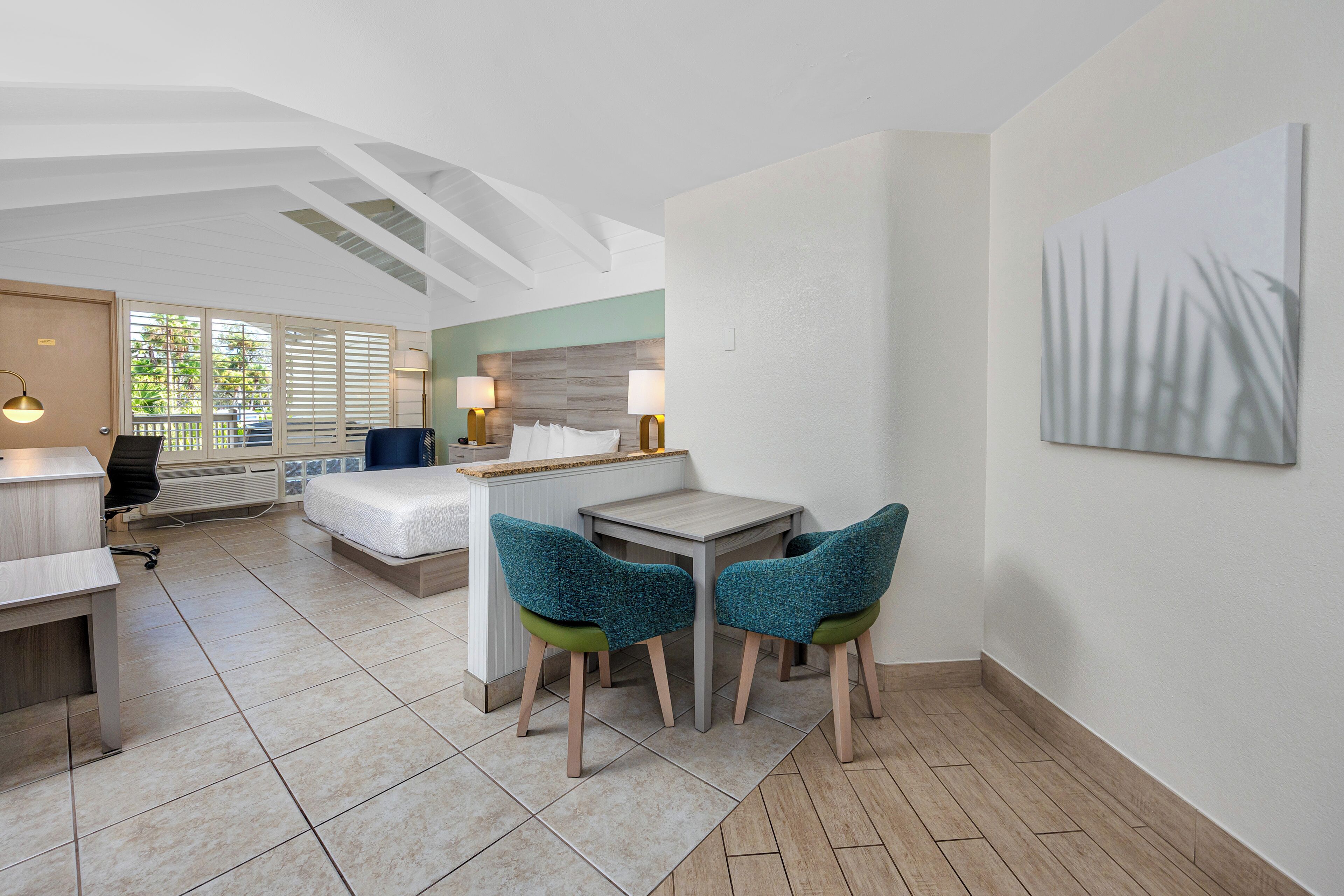 Una habitación de hotel moderna con una cama, un escritorio, sillas, una mesita pequeña y un cuadro en la pared.