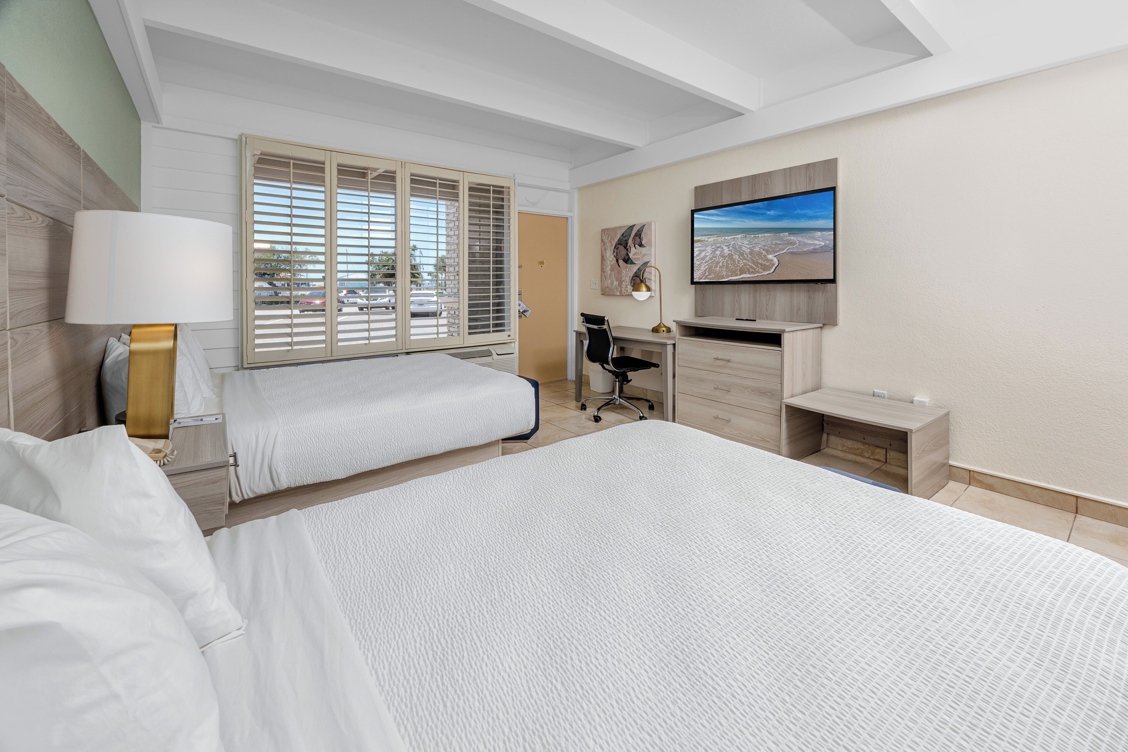 Quarto de hotel com duas camas, uma mesa, uma cadeira, uma televisão e janela com persianas.