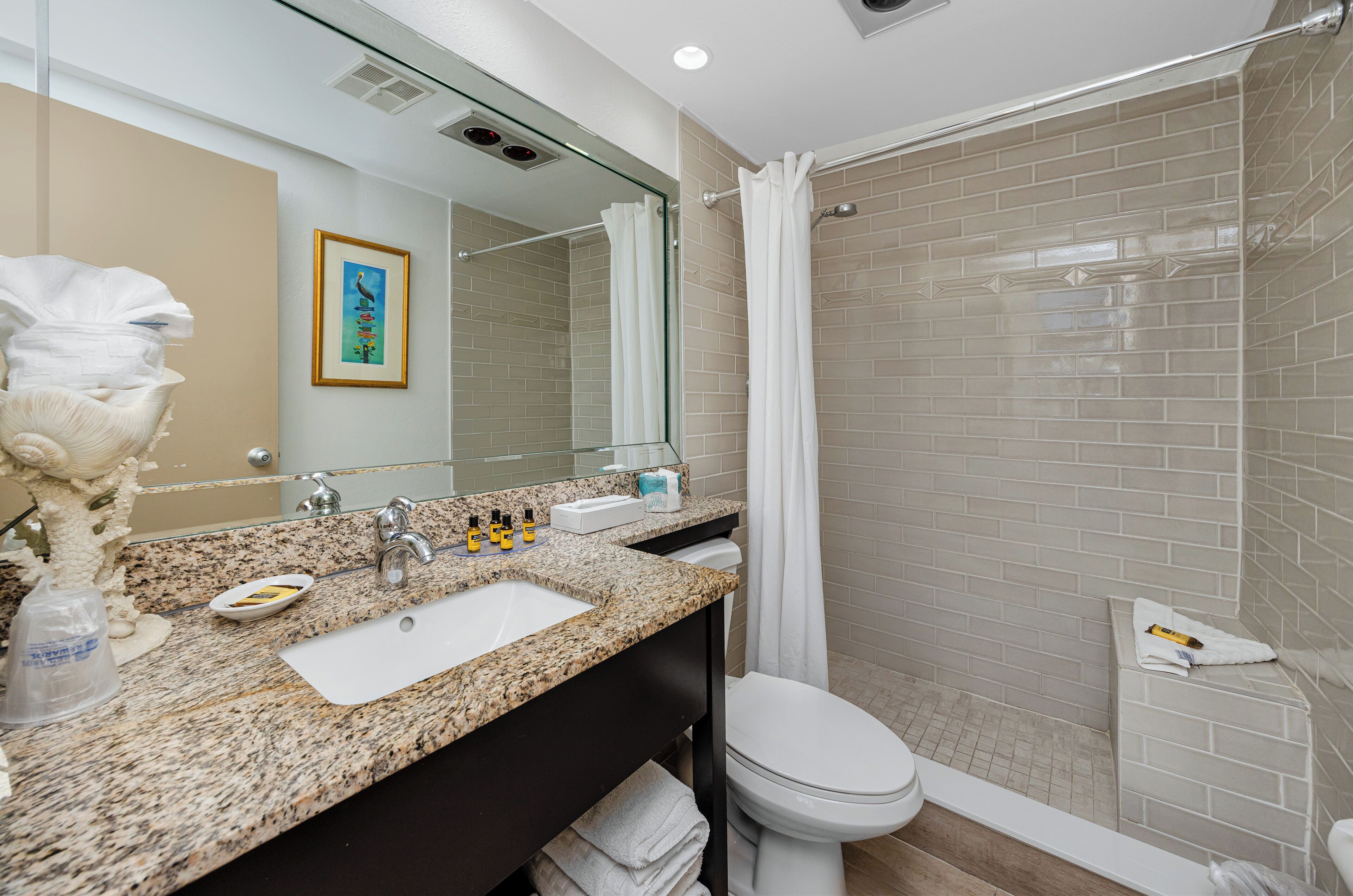 Un baño moderno con encimera de granito, un lavabo blanco, un inodoro y cabina de ducha con mampara de vidrio.