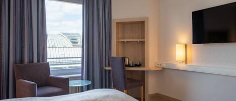 Habitación de hotel con cama, dos sillones, una mesita redonda, un escritorio con silla, televisor montado en la pared y ventana con cortinas.