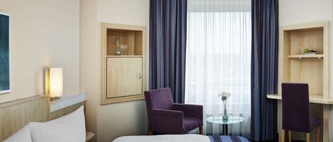 A hotel room with a bed, two chairs, a small table with a vase, and a window with curtains.