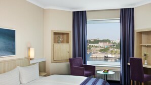 Habitación de hotel con cama, dos sillas, una mesita con fruta, un armario y un ventanal con vistas a la ciudad.