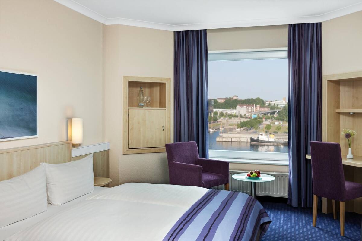 Een hotelkamer met een bed, twee stoelen, een klein tafeltje met fruit, een kast en een groot raam met uitzicht over de stad.