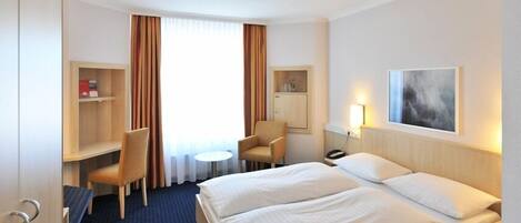 A hotel room with a bed, two chairs, a desk, a small table, a nightstand, and a painting on the wall.