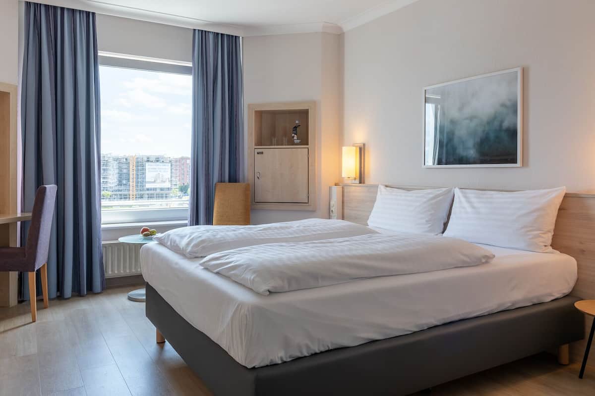 Een hotelkamer met een groot bed, een stoel, een bureau, een lamp en uitzicht op de stad.