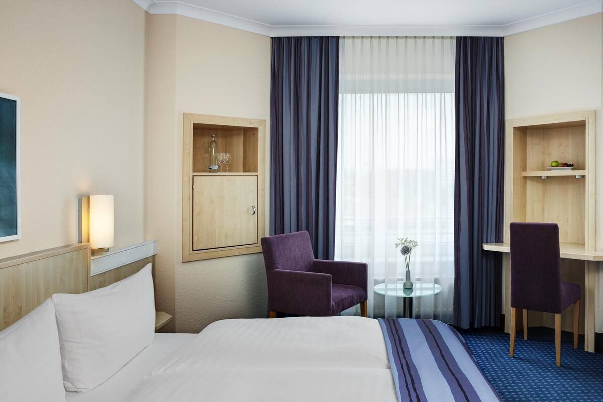 Hotelkamer met een bed, twee stoelen, een klein tafeltje, een kastje met een fles en glazen, en een groot raam met gordijnen.
