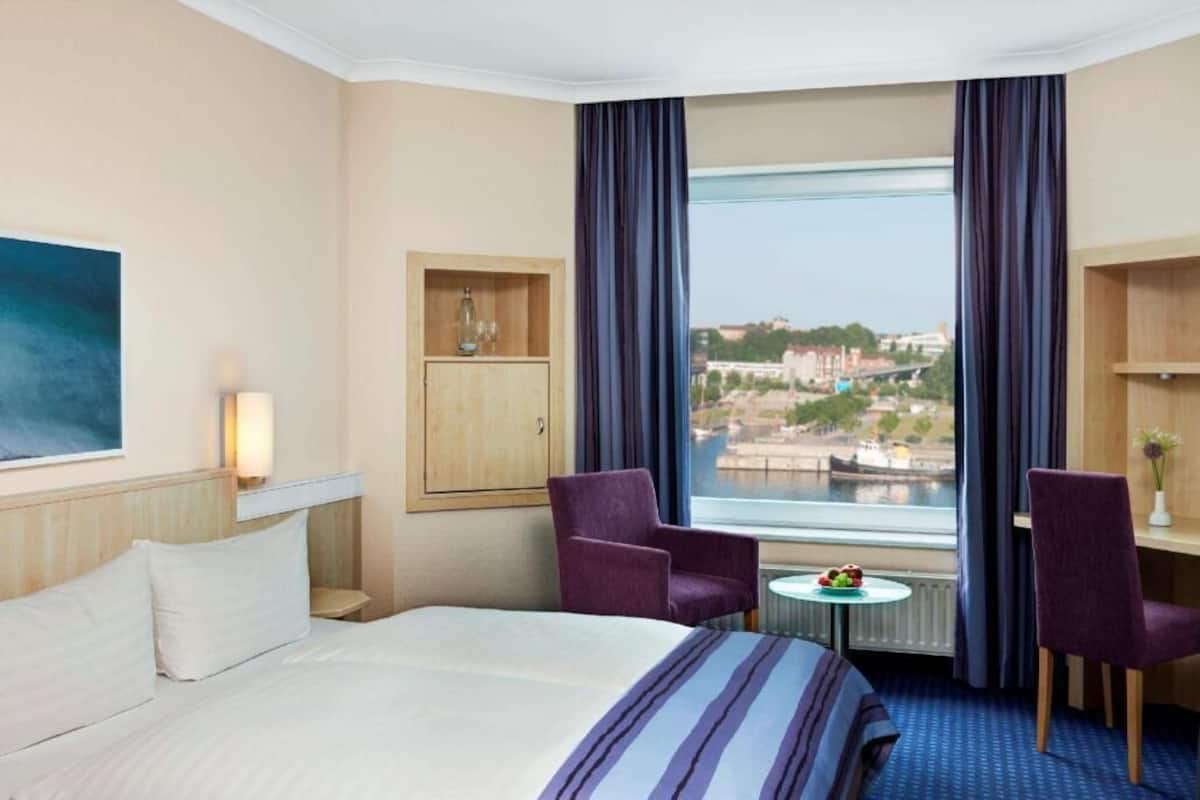 Hotelkamer met een bed, twee stoelen, een tafeltje, een kast en een raam met uitzicht op een stadsgezicht.
