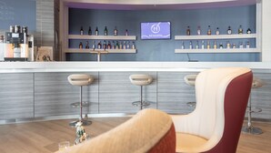 Zona de bar con barra, estantes llenos de botellas de licor, una pantalla de televisión mostrando 'REWARDS' y tres taburetes de bar.