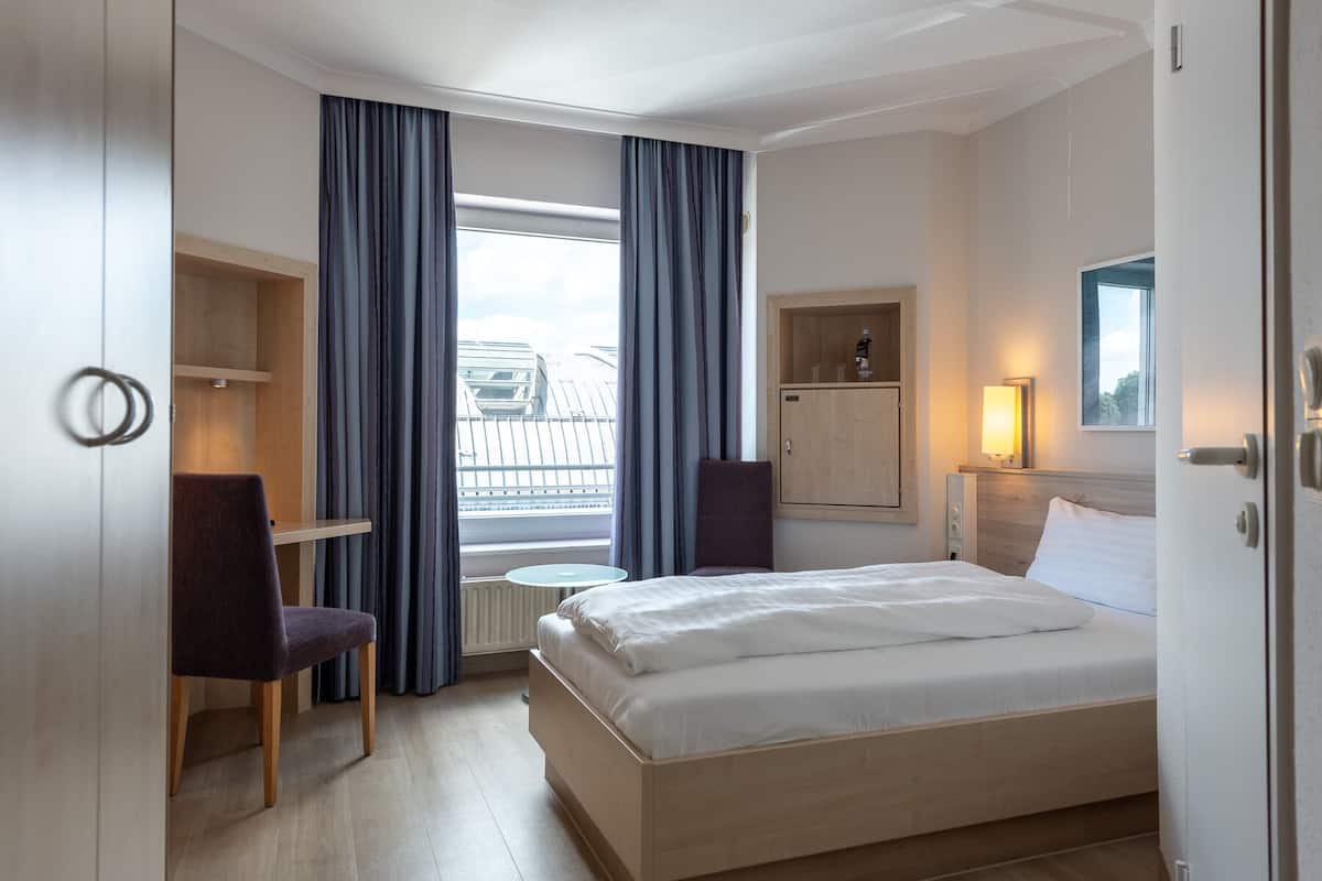 Een hotelkamer met een bed, een bureau, een stoel, een klein tafeltje, een kast en een raam met gordijnen.