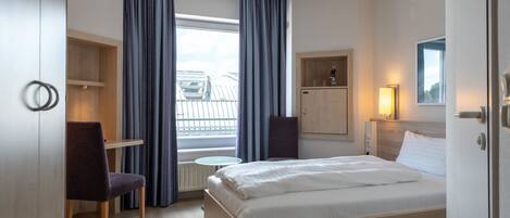A hotel room with a bed, a desk, a chair, a small table, a cabinet, and a window with curtains.