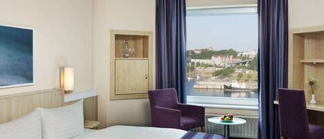 A hotel room with a large bed, two armchairs, a small table with fruit, a cabinet, and a view of a cityscape through a window.
