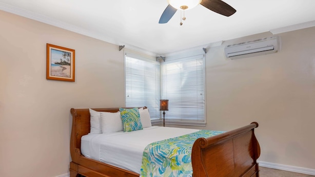 A bedroom with a wooden bed, white bedding, a beach-themed picture on the wall, and a ceiling fan.