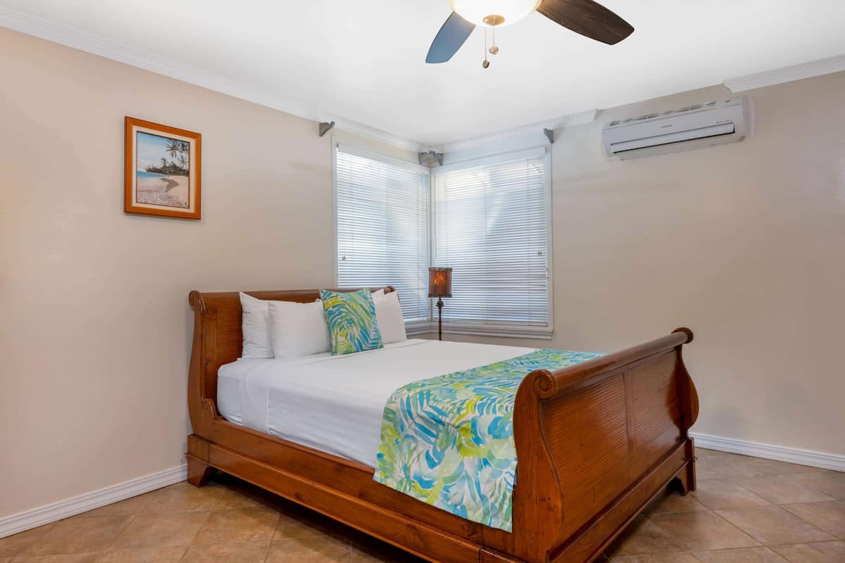 A bedroom with a wooden bed, white bedding, a beach-themed picture on the wall, and a ceiling fan.