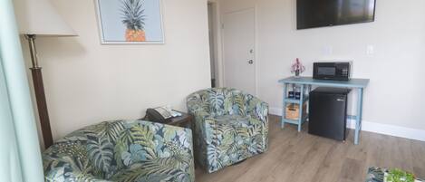 Una sala de estar con dos sofás estampados, un televisor de pantalla plana, un microondas y un refrigerador pequeño.
