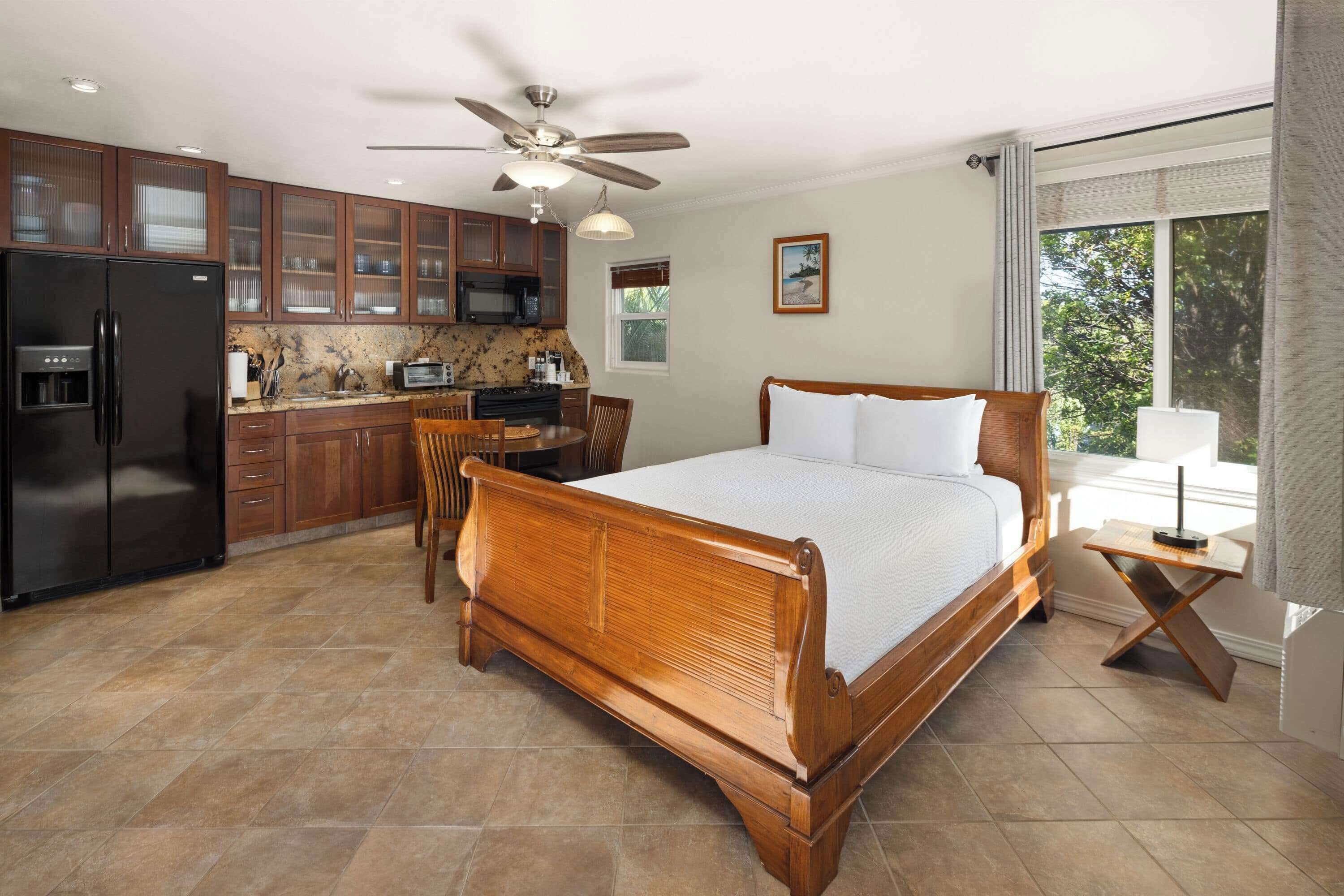 A bedroom with a wooden bed, a dining table with chairs, a kitchen with cabinets and appliances, and a ceiling fan.