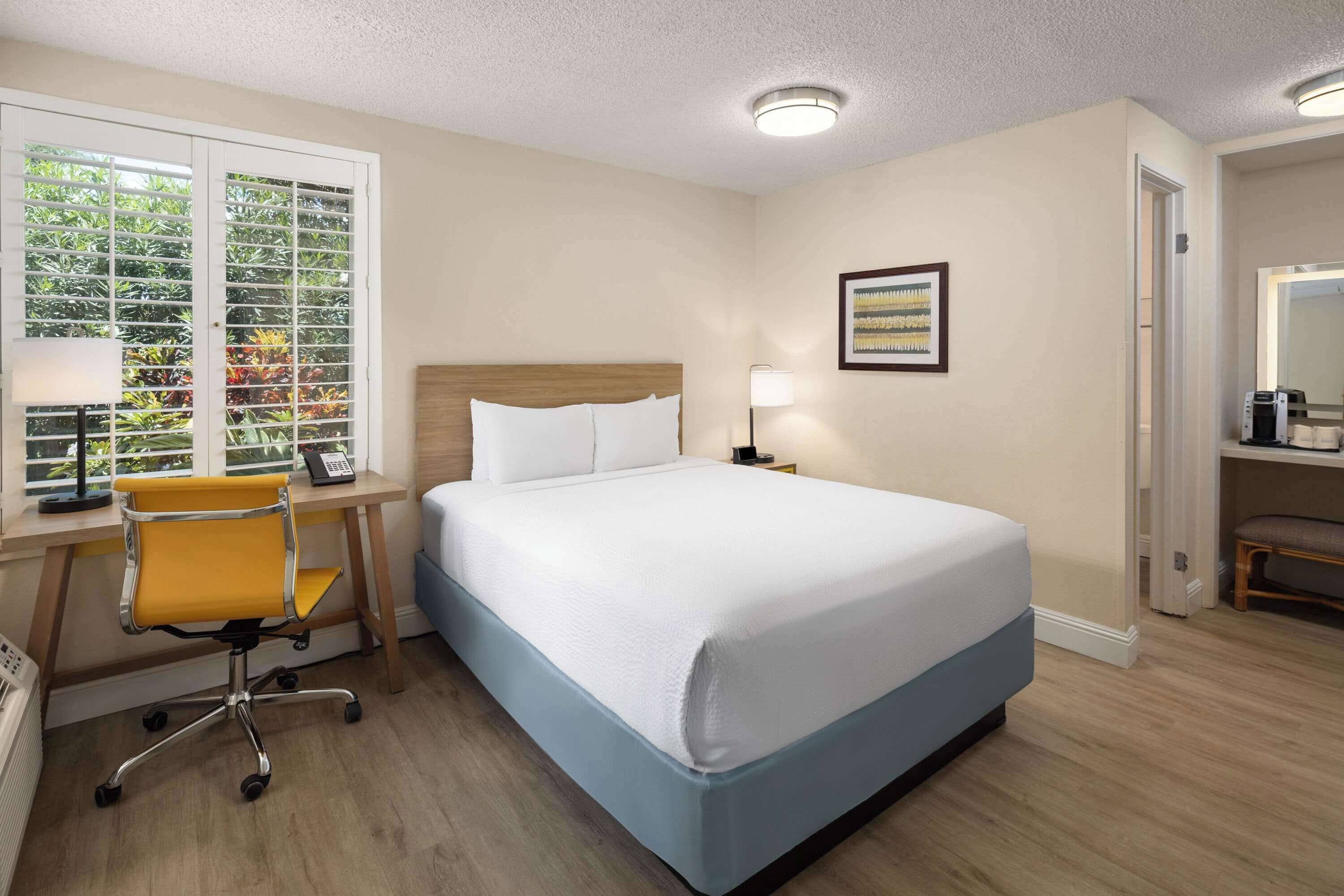 A hotel room with a bed, desk, chair, and a window with blinds.