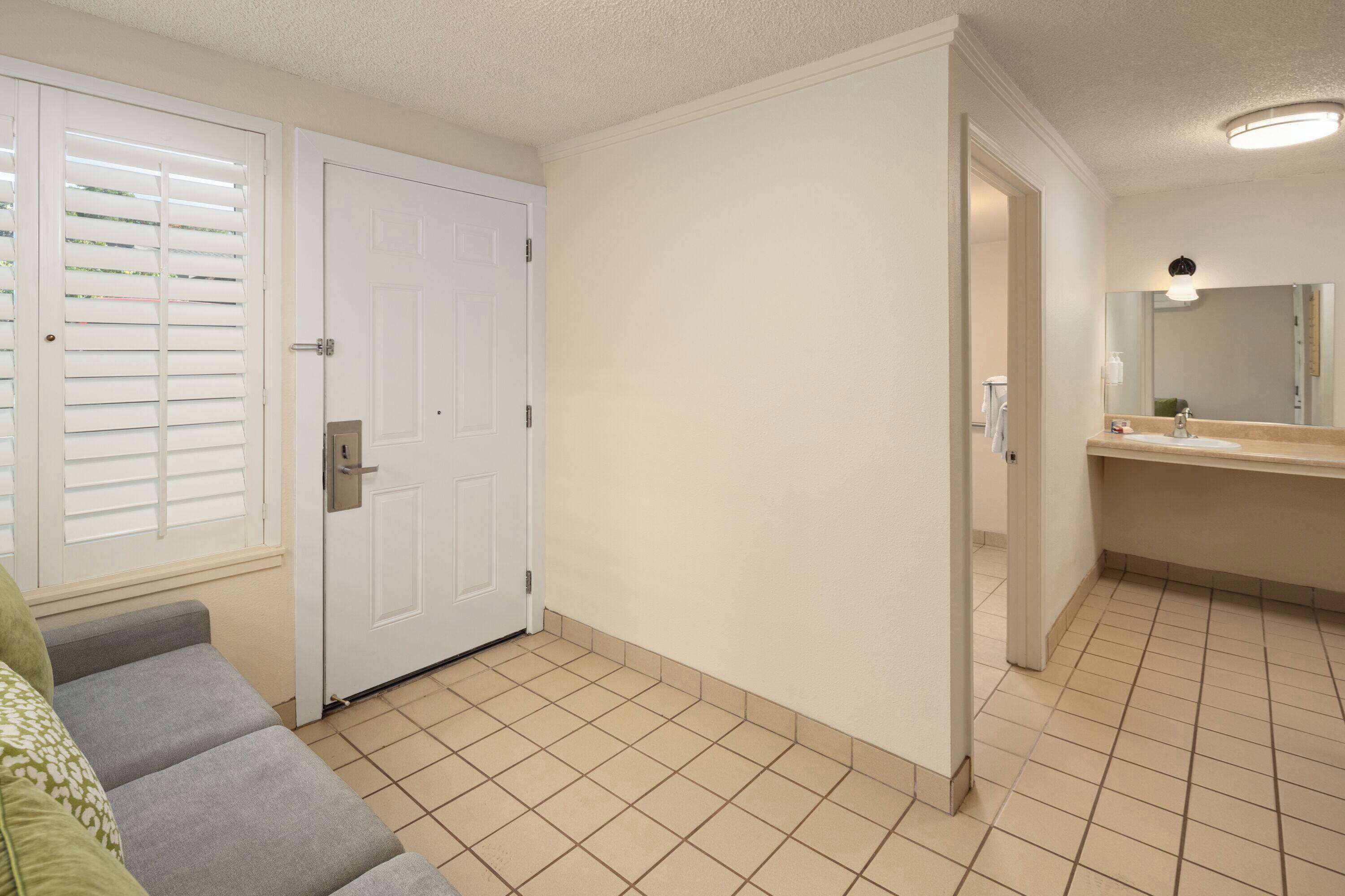 A clean, well-lit hallway with a white door, a small seating area with a green cushion, and a bathroom with a sink and mirror.