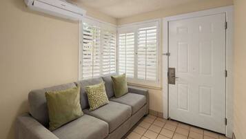 A room with a grey sofa, two green cushions, a white door, and a window with white blinds.