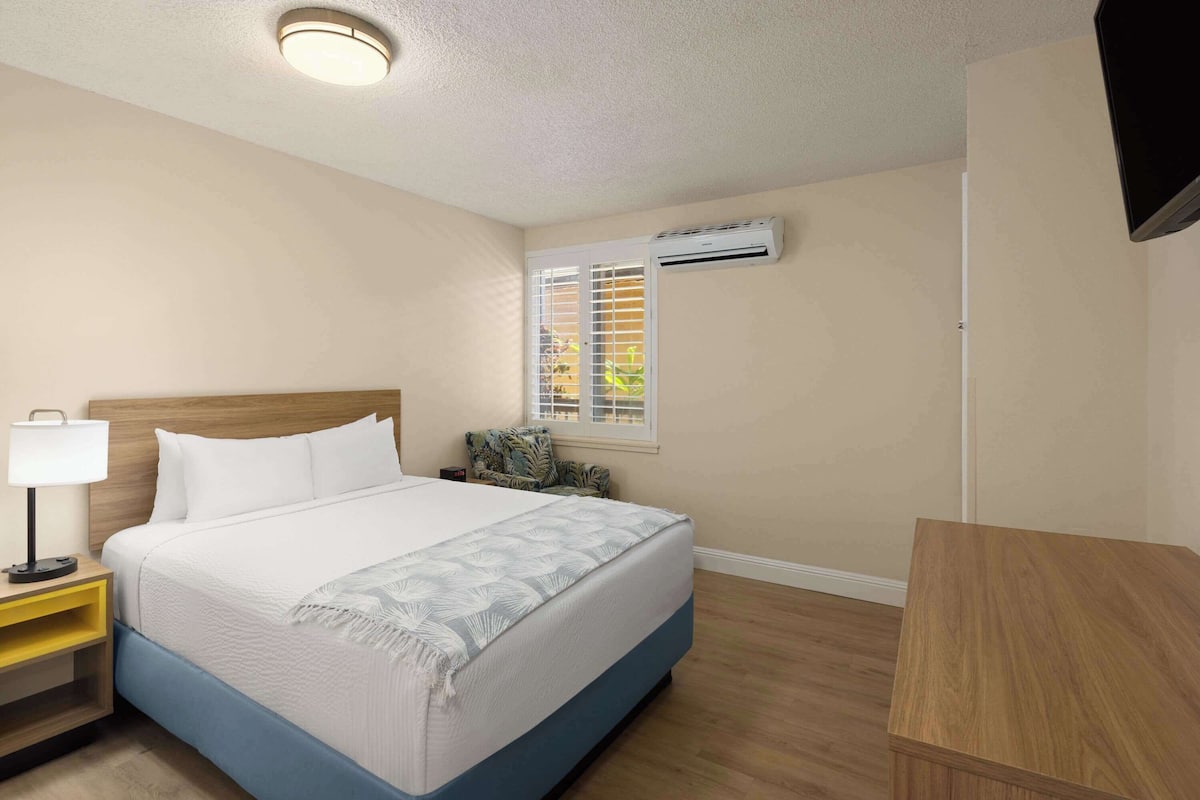 A neatly arranged hotel room with a bed, bedside table, lamp, air conditioning unit, window with blinds, and a small wooden table.