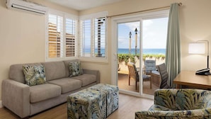 A living room with a sofa, ottoman, and chairs, a table with a lamp, and a view of the beach through a sliding glass door.