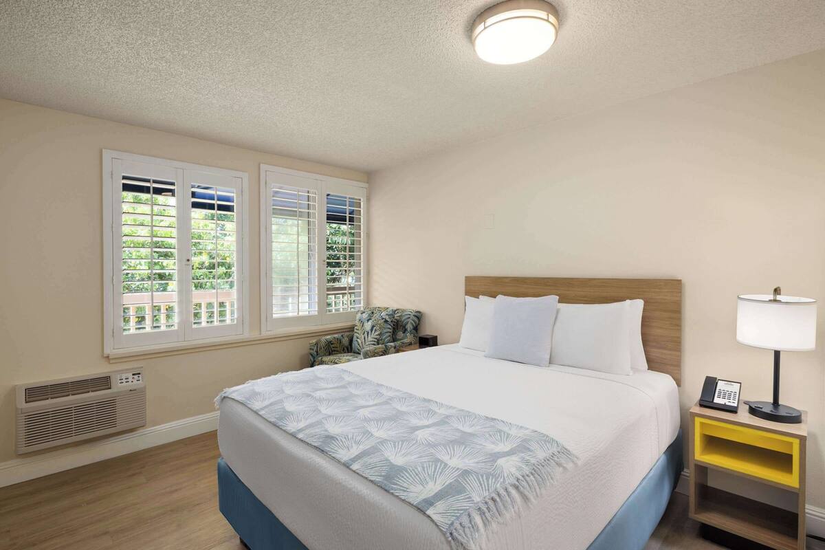A hotel room with a bed, a nightstand, a chair, and a window with blinds.