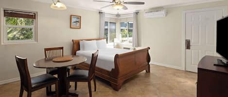 A bedroom with a bed, a desk with chairs, a ceiling fan, a window with blinds, and a door.