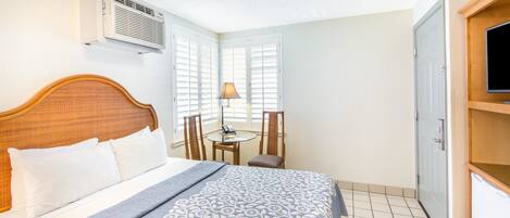 A hotel room with a bed, a desk with a lamp, a TV, and an air conditioning unit.