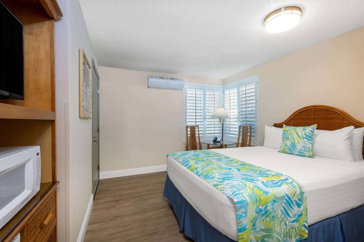 A hotel room with a bed, a television, an air conditioning unit, and a window with blinds.