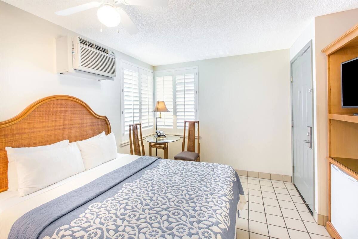 A hotel room with a bed, a desk, a chair, a television, and an air conditioning unit.