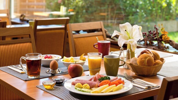 A breakfast table with a variety of food items, drinks, and a vase with a white flower. - Leonardo Hotel Karlsruhe (Karlsruhe)