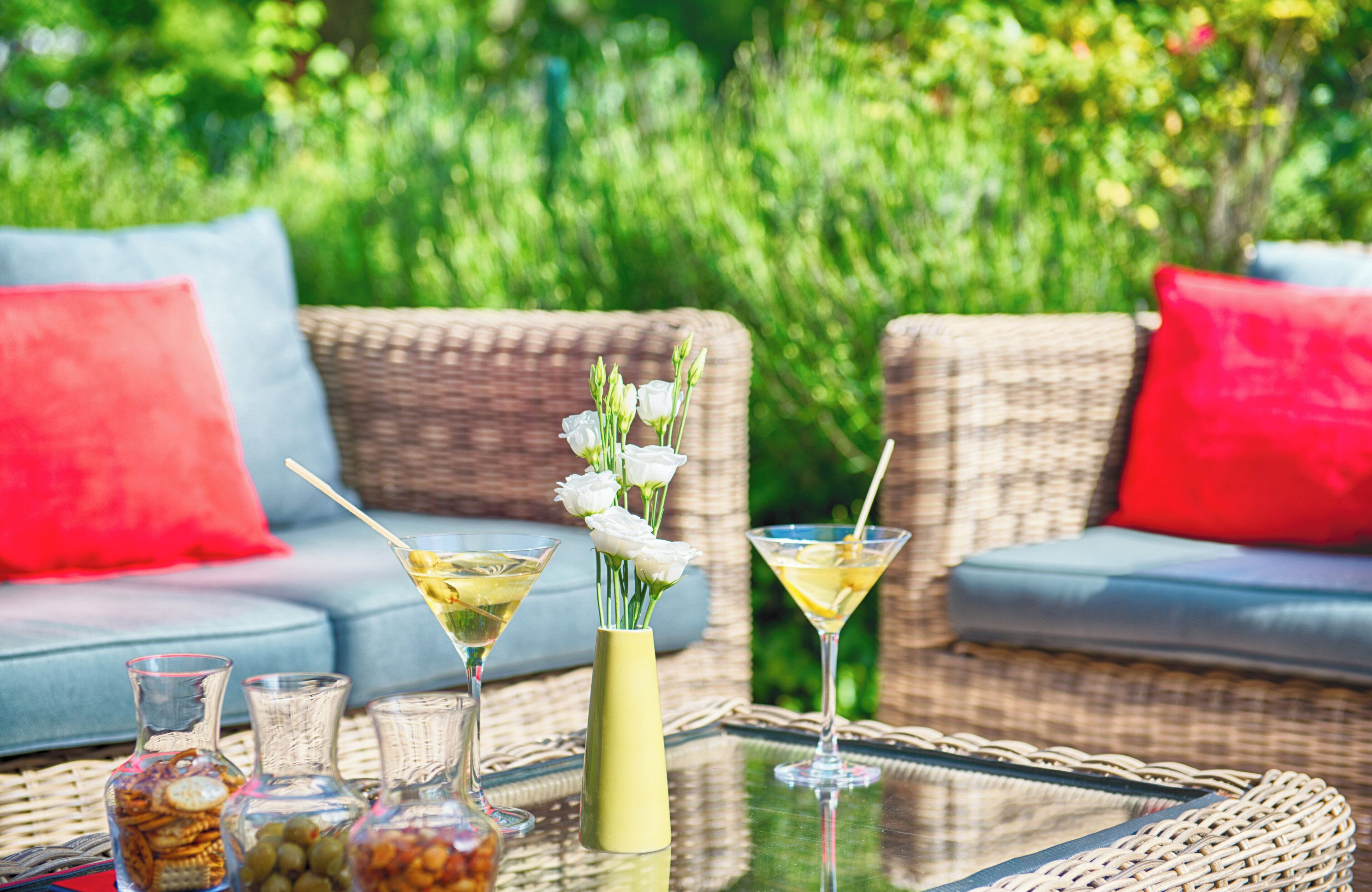 A wicker sofa with red cushions, a glass table with drinks and condiments, and a vase with white flowers.