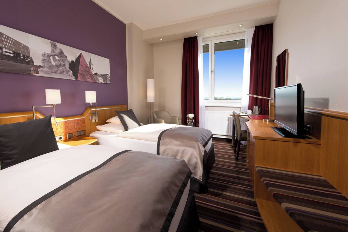 A hotel room with two beds, a television, a desk, and a view of the city.