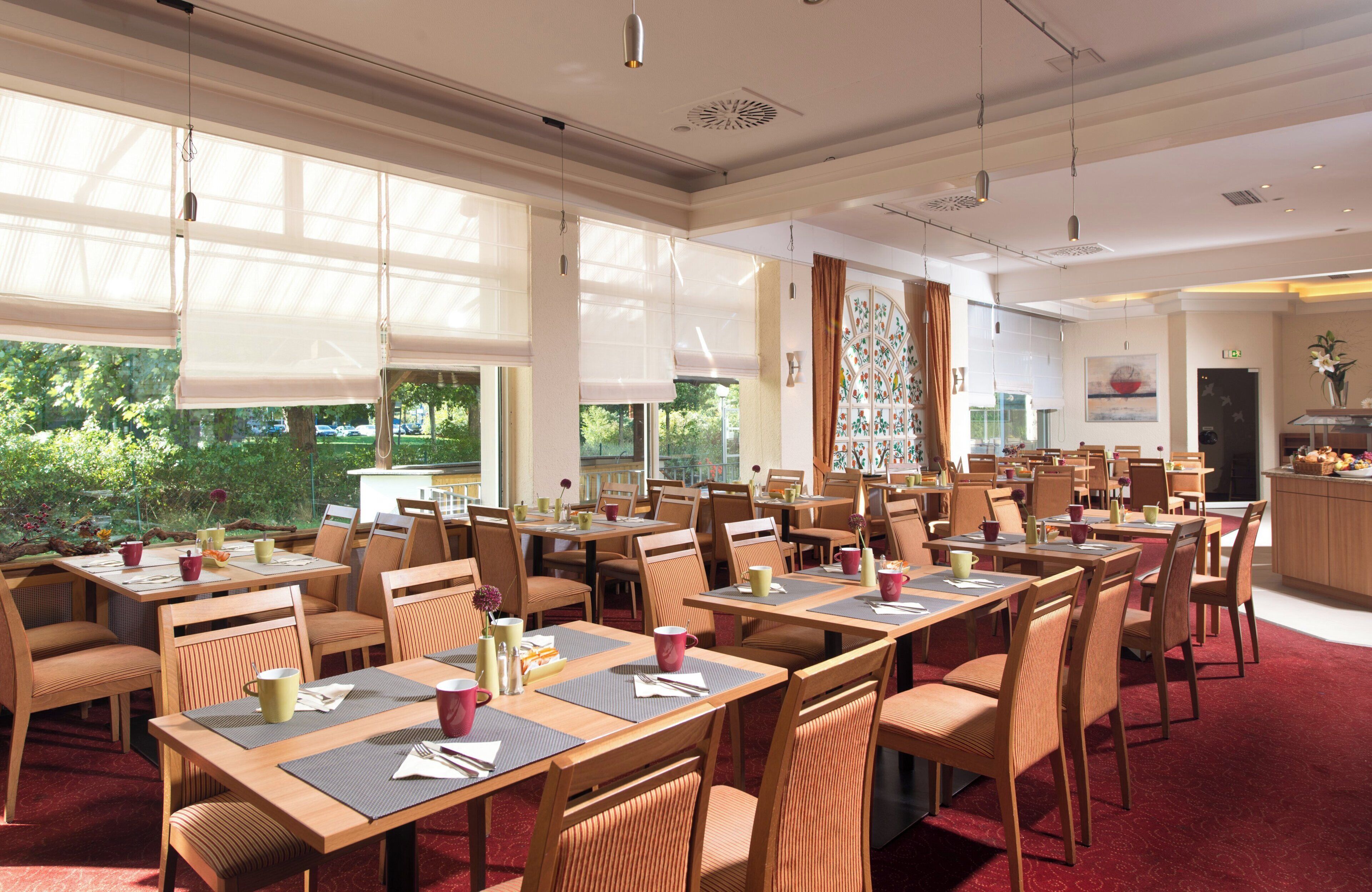 A spacious dining area with multiple tables and chairs, large windows with blinds, and a view of greenery outside.