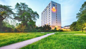 A modern hotel building with a prominent sign, surrounded by lush greenery and a pathway. - Leonardo Hotel Karlsruhe (Karlsruhe)