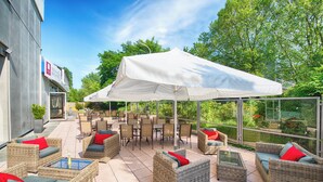 An outdoor patio area with wicker furniture, a white canopy, and a glass enclosure overlooking a garden. - Leonardo Hotel Karlsruhe (Karlsruhe)