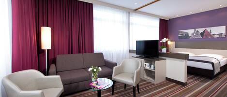 A hotel room with a sofa, armchair, TV, bed, and a small table with a vase of flowers.
