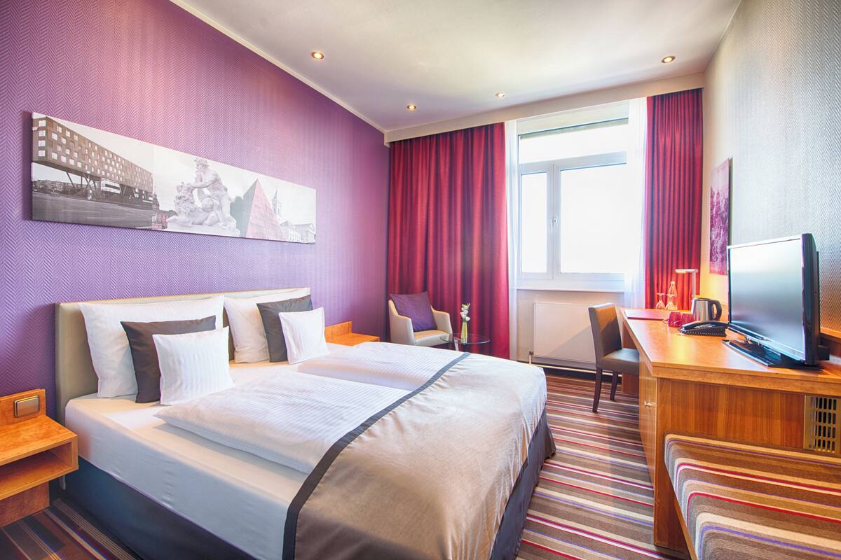 A hotel room with a large bed, a desk with a TV, a chair, and a window with curtains.