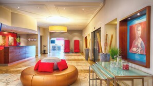 A hotel lobby with a reception desk, a round seating area with red cushions, a glass table with decorative items, and a large framed artwork on the wall. - Leonardo Hotel Karlsruhe (Karlsruhe)