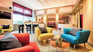 A spacious living area with a yellow armchair, a blue armchair, and a red sofa. There is a small coffee table with a vase and a lamp. The room has a large window with a red curtain and a flat-screen TV mounted on the wall. - Leonardo Hotel Karlsruhe (Karlsruhe)