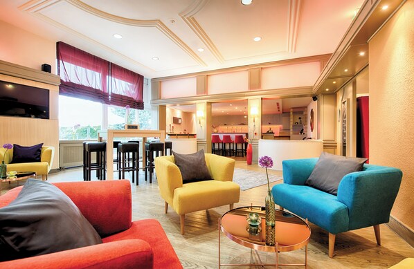 Un salón espacioso con un sillón amarillo, un sillón azul y un sofá rojo. Hay una mesita de centro con un jarrón y una lámpara. La habitación tiene un ventanal grande con una cortina roja y un televisor de pantalla plana montado en la pared.