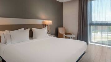 Habitación de hotel con una cama grande, una mesita de noche, una lámpara, una silla y vistas a través de cortinas translúcidas.