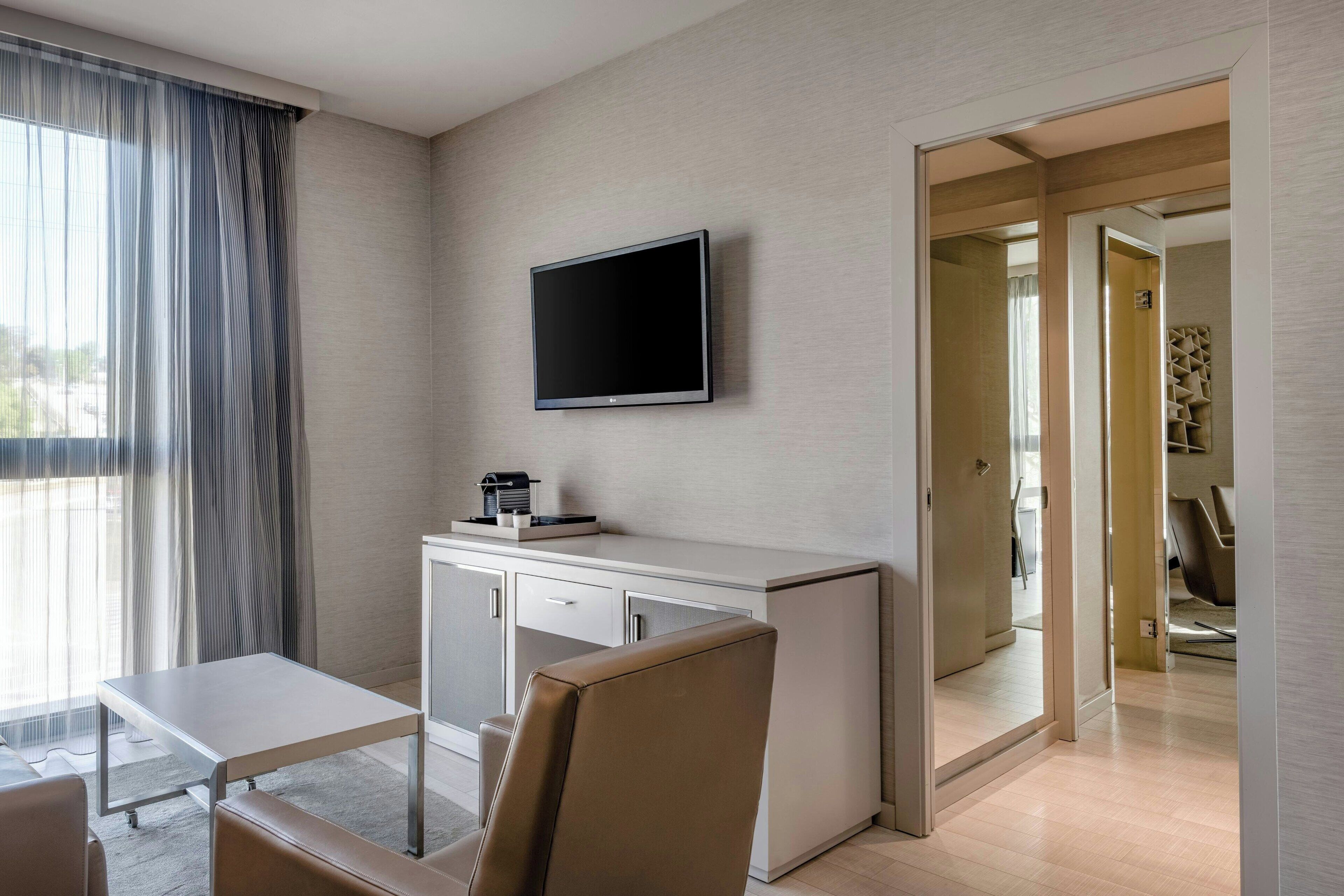 A modern hotel room with a flat-screen TV mounted on the wall, a small desk with a coffee maker, a cushioned chair, and a large window with sheer curtains.