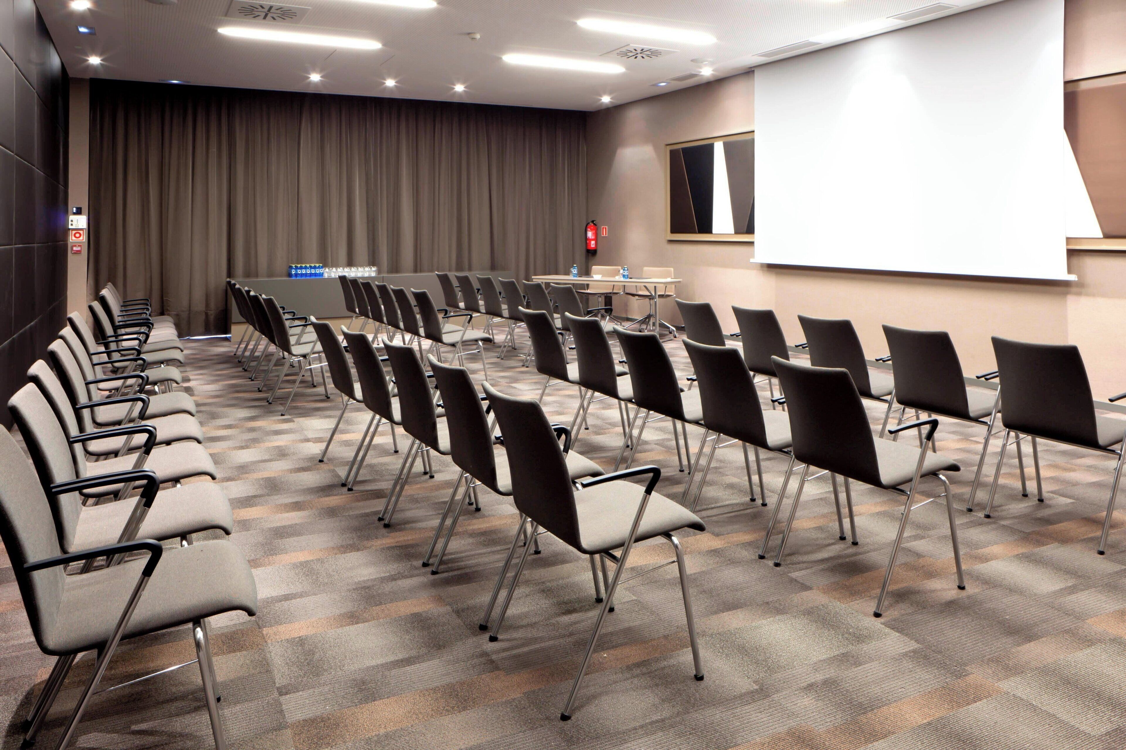 A conference room with rows of chairs, a large table, a projector screen, and a fire extinguisher.