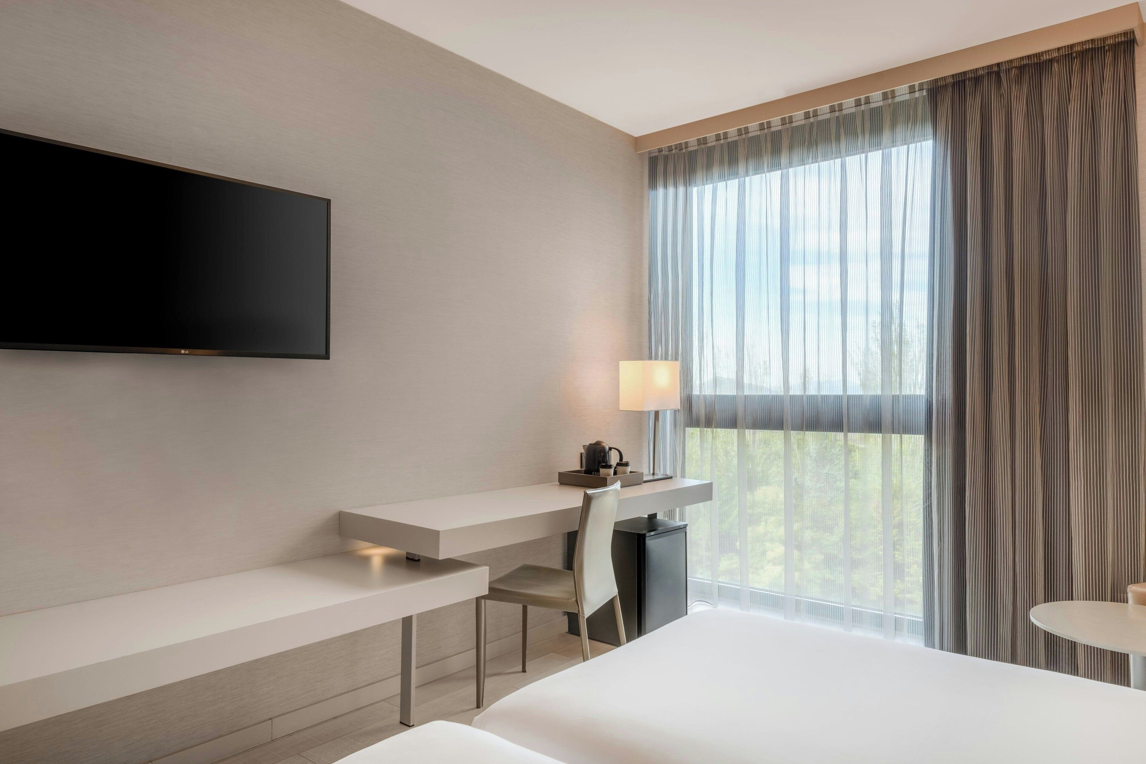Ein modernes Hotelzimmer mit einem wandmontierten Flachbildfernseher, einem Schreibtisch mit Stuhl, einem kleinen Tisch und einem Bett.