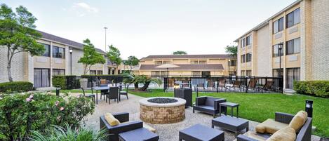 An outdoor patio area with seating arrangements, a fire pit, and a building in the background.