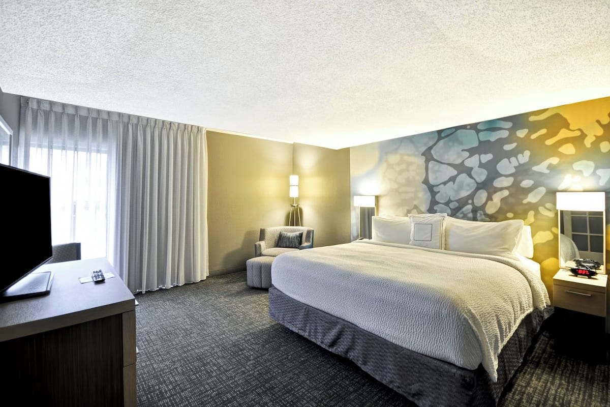 A hotel room with a large bed, a desk with a TV, a chair, and a lamp.