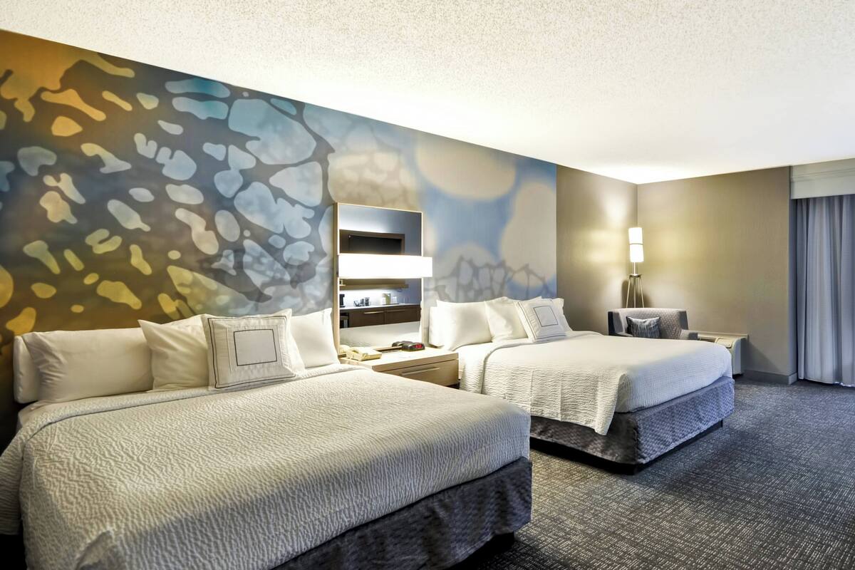 A hotel room with two beds, a wall-mounted TV, a desk, and a lamp.